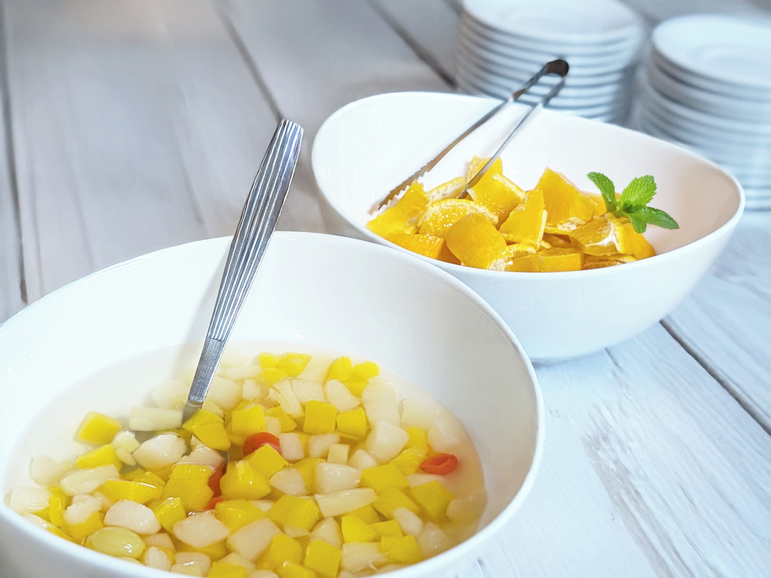 Two white bowls on a wooden table, one containing diced yellow fruit and the other with orange slices and mint leaves.