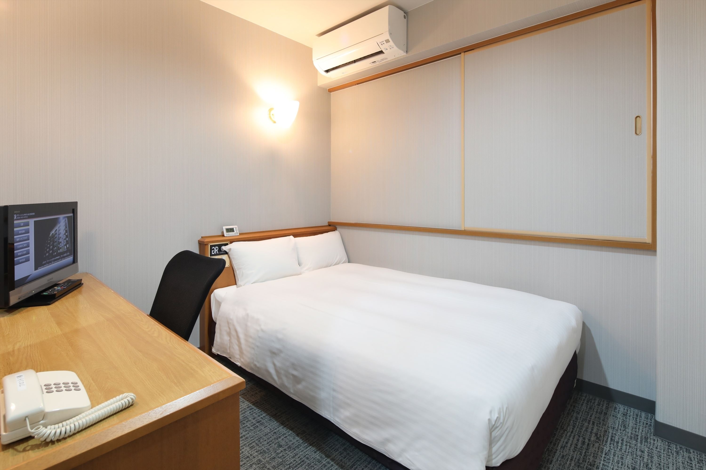 Quarto de hotel com cama, escrivaninha com telefone, cadeira, ar condicionado e luminária na parede.