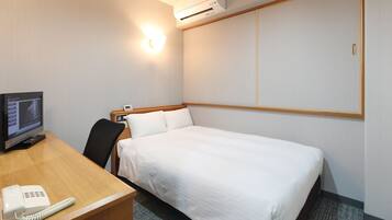 Quarto de hotel com cama, escrivaninha com telefone, cadeira, ar condicionado e luminária na parede.