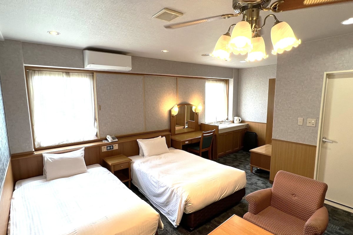 A hotel room with two beds, a desk, a chair, a small table, a mirror, and a ceiling fan.