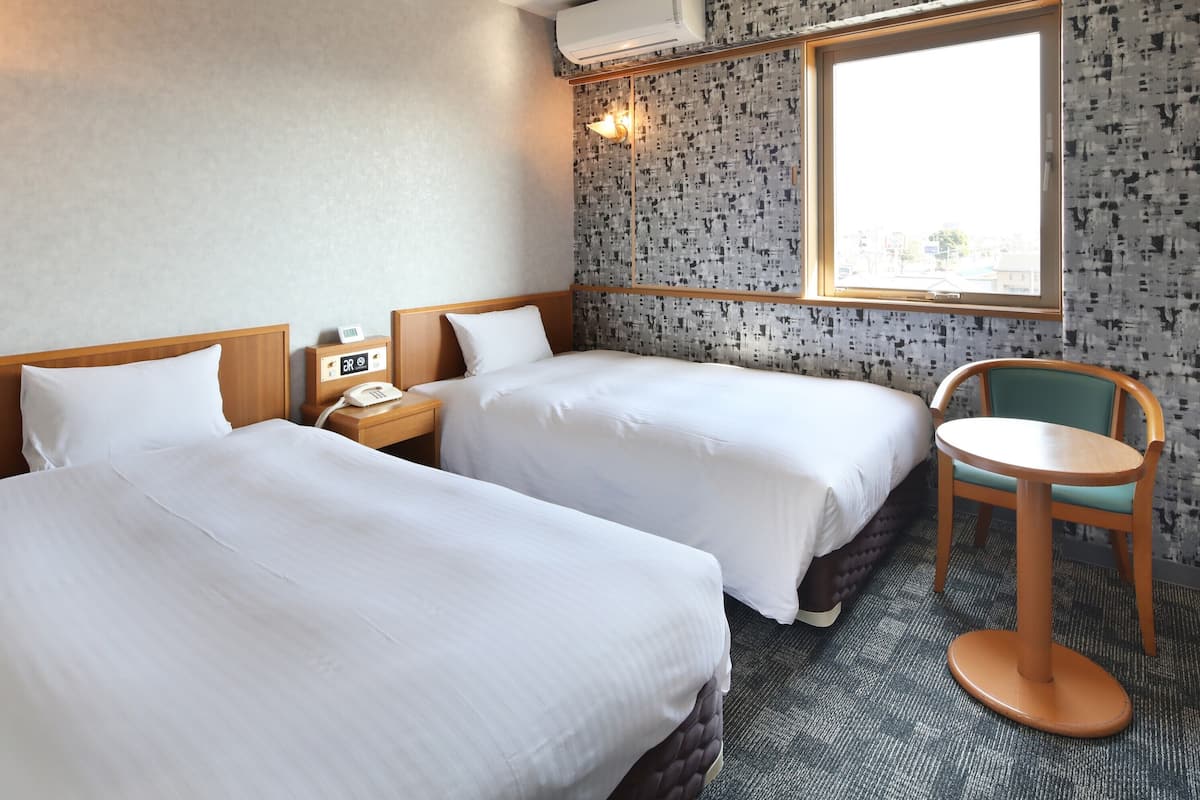 A hotel room with two beds, a small table, a chair, a bedside table with a phone, and a window with a view.