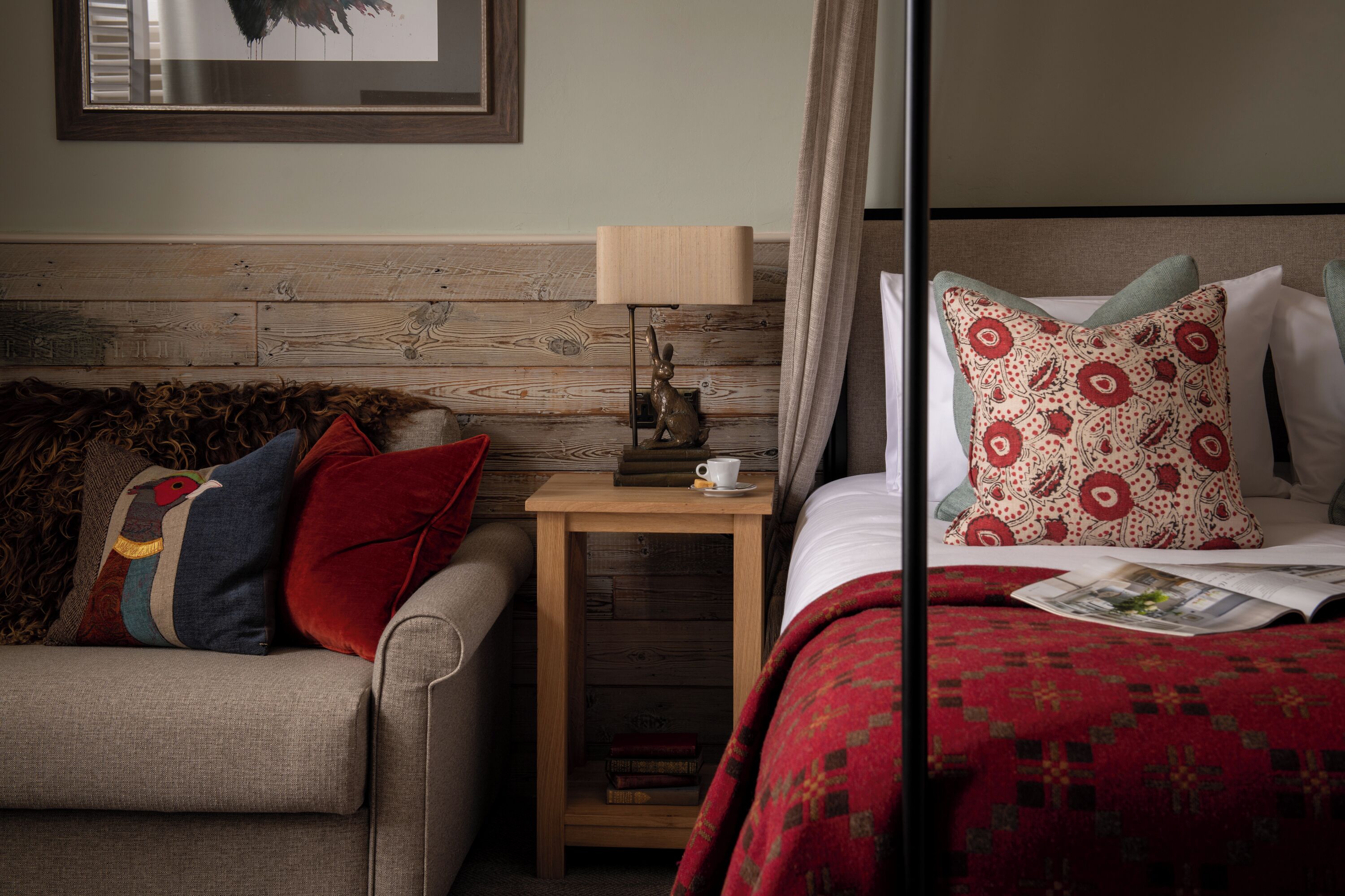 Habitación de hotel con cabecera de madera, una pequeña mesita de noche con lámpara, edredón estampado rojo y un sofá gris con varios cojines.