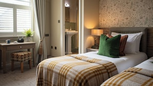 A hotel room with two beds, a desk, a chair, a window with curtains, a bathroom, and a wall with a floral pattern. - The Golden Lion (Settle)