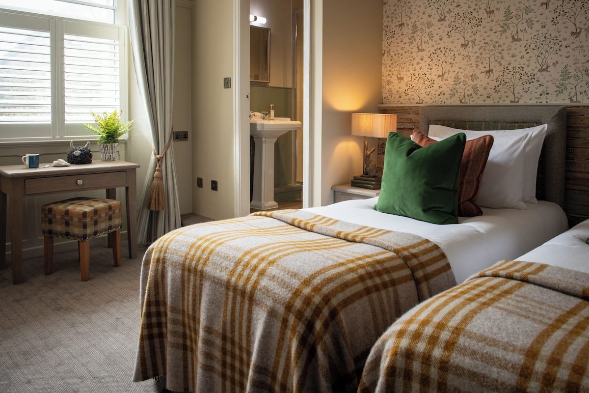 A hotel room with two beds, a desk, a chair, a window with curtains, a bathroom, and a wall with a floral pattern.