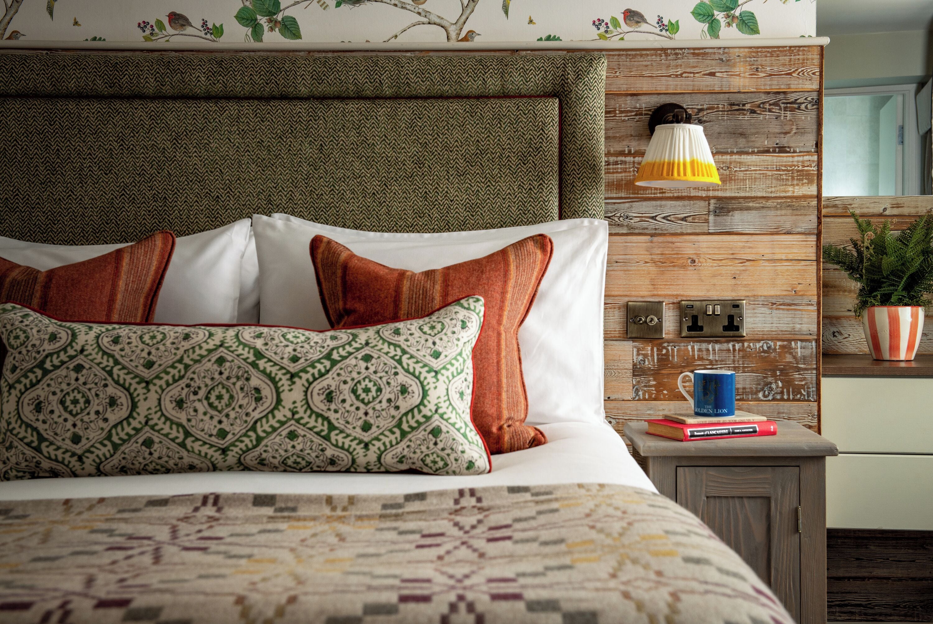 Una cama bien hecha con ropa de cama y almohadas estampadas, una mesita de noche con libros y una taza, y una lámpara fijada a la pared.