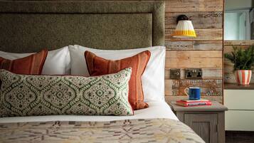 Una cama bien hecha con ropa de cama y almohadas estampadas, una mesita de noche con libros y una taza, y una lámpara fijada a la pared.