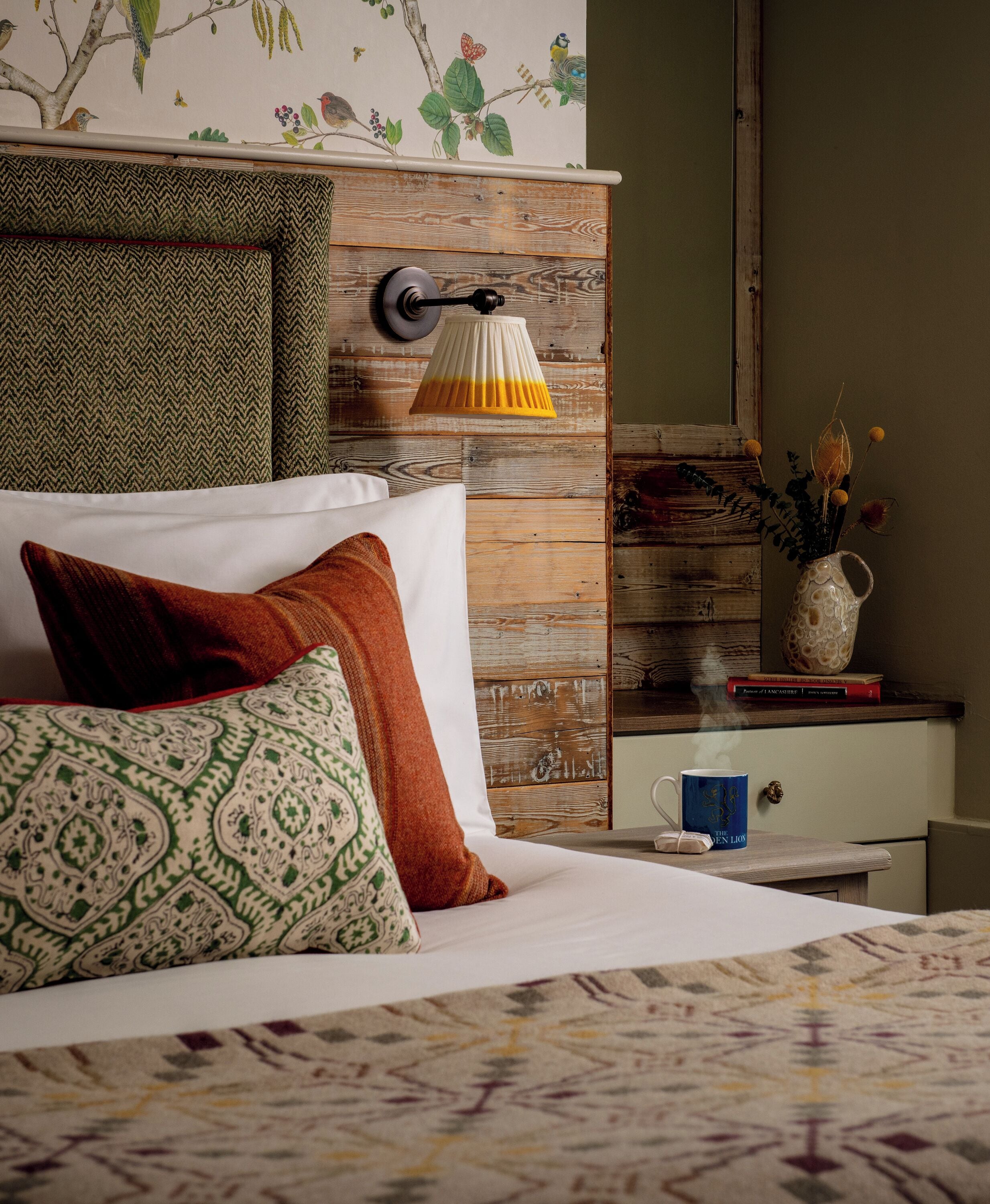 A neatly made bed with patterned pillows, a wooden headboard, a bedside table with a lamp, a vase with flowers, and a book.