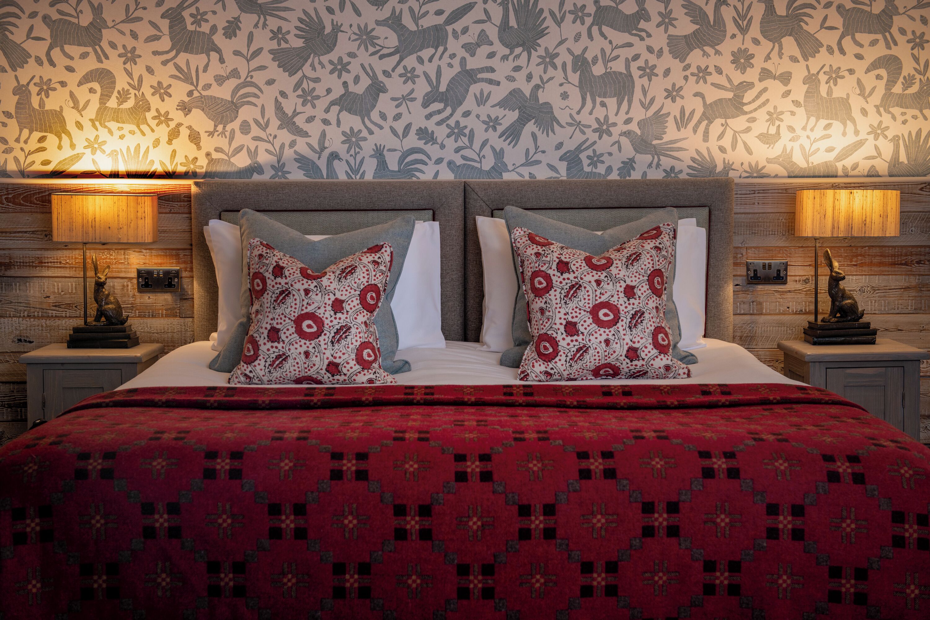 A bed with a red patterned bedspread, two pillows with floral designs, and a wall with a floral wallpaper pattern.
