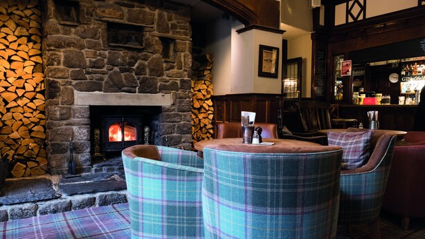 A cozy pub with a stone fireplace, plaid furniture, and a bar area. - The Golden Lion (Settle)