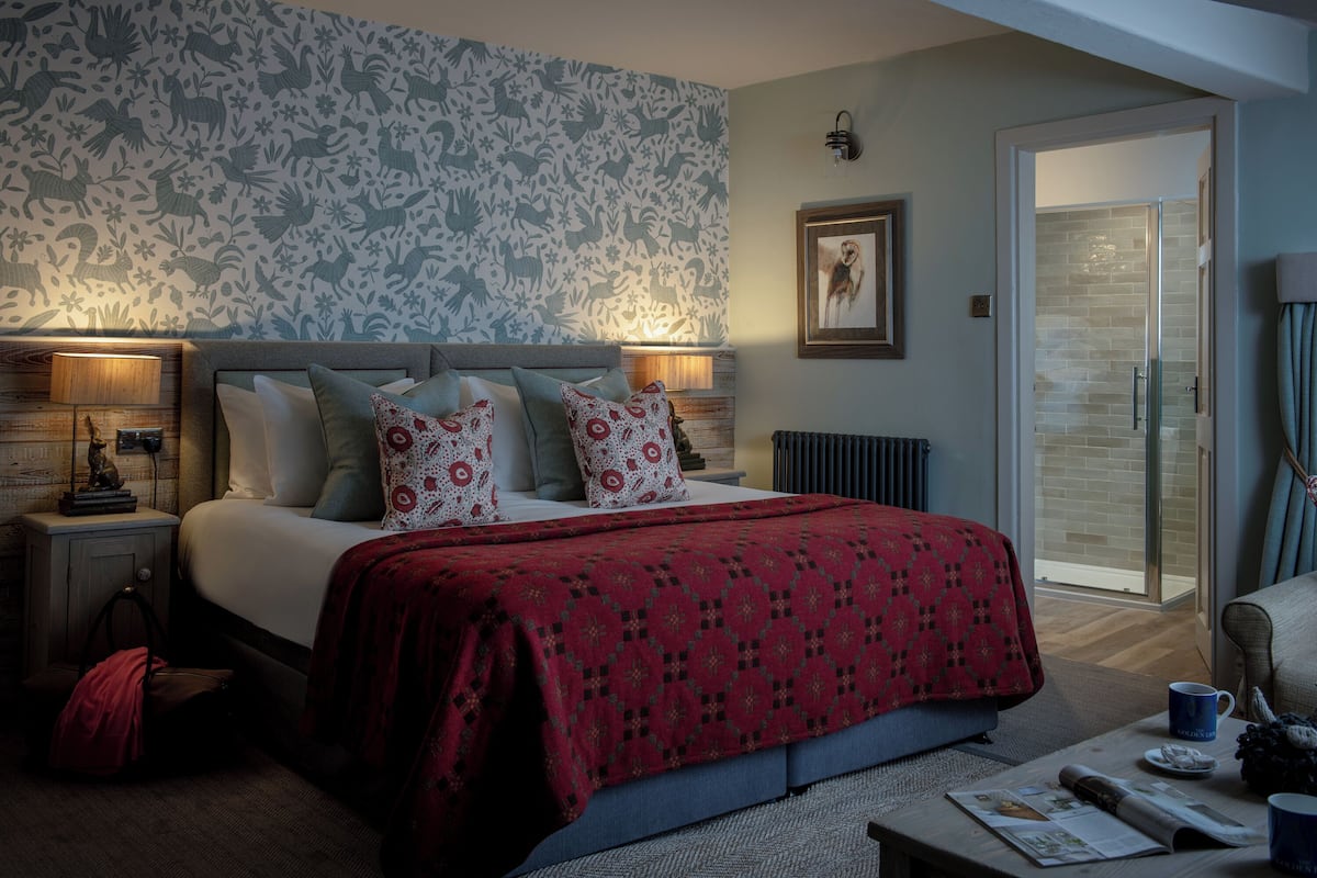 A bedroom with a bed, bedside tables, a nightstand, a red patterned bedspread, a wall with floral wallpaper, a framed picture, and a glass door leading to a bathroom.