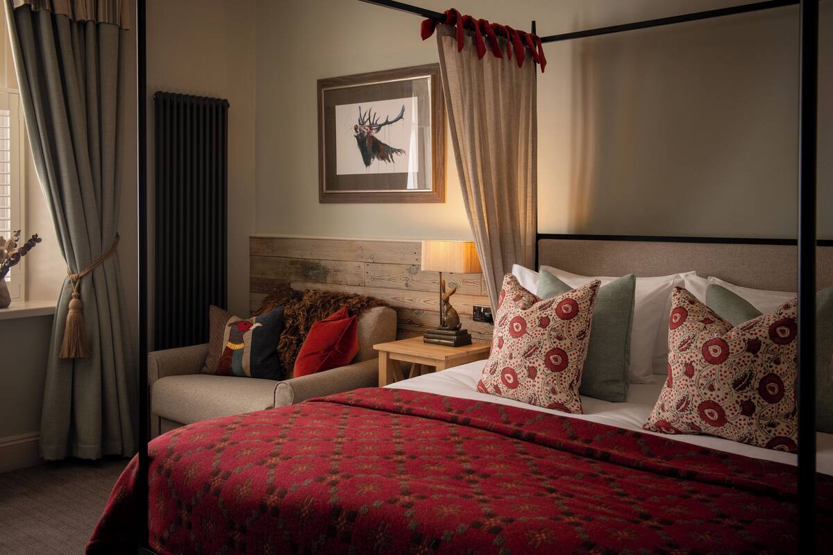 A bedroom with a four-poster bed, a wooden headboard, a nightstand with a lamp, and a framed deer head on the wall.