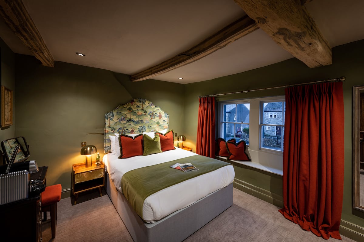 A bedroom with a large bed, a window with red curtains, a nightstand with a lamp, and a view of the outside.