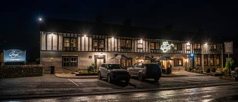 Un edificio de dos pisos con un letrero que dice "The Lodge". Hay dos coches estacionados frente a él, y el edificio está iluminado por luces exteriores.