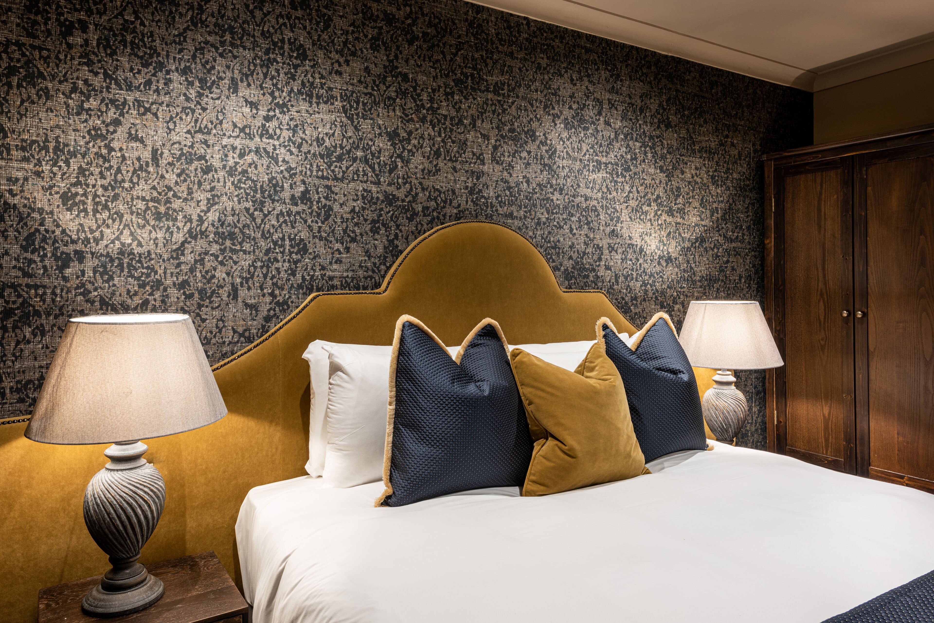 A neatly made bed with a headboard, two bedside lamps, and a patterned wall in the background.