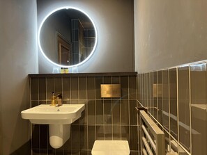 A modern bathroom with a white sink, a mirror with a circular light, and dark tiled walls.