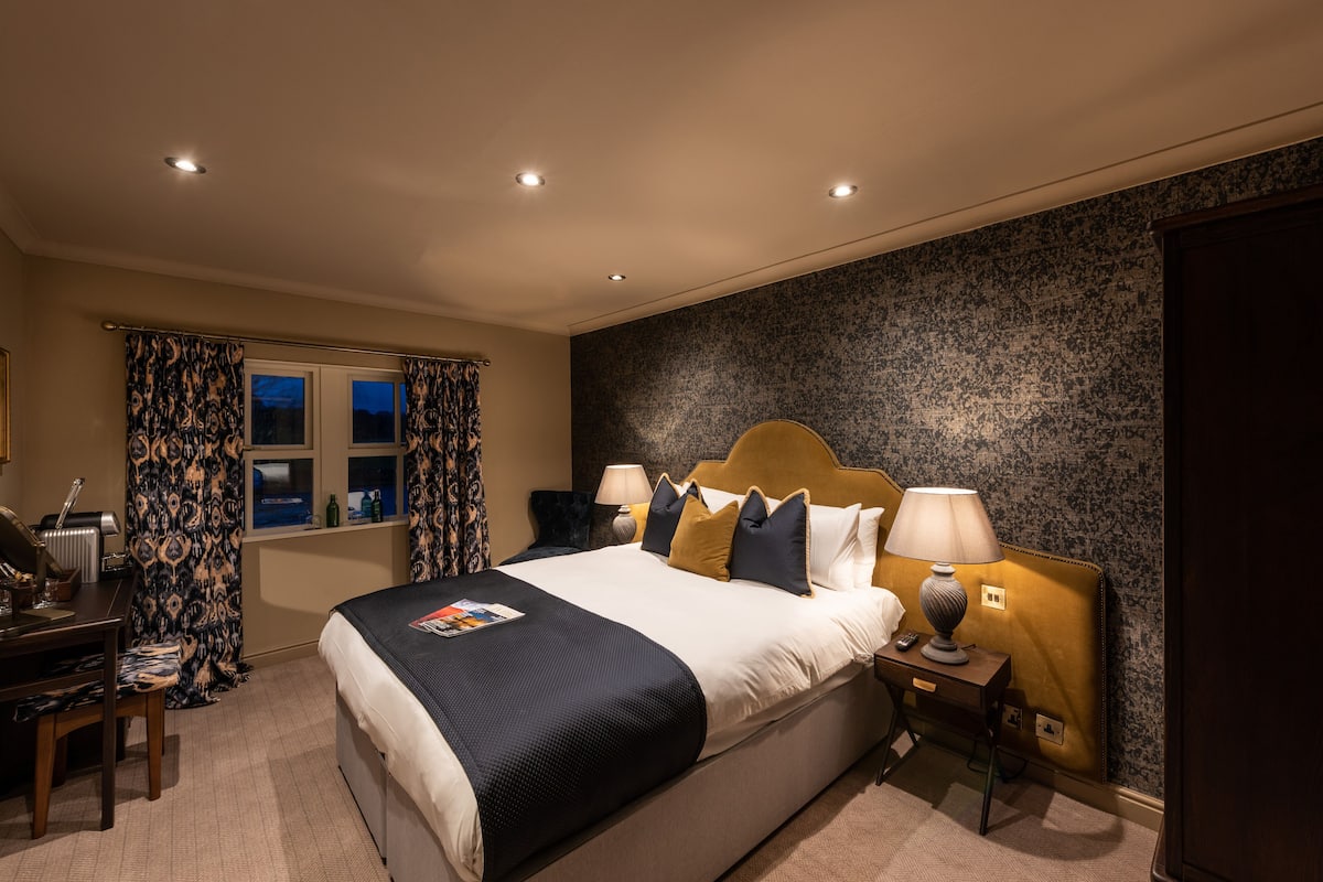 A hotel room with a large bed, two bedside tables with lamps, a desk with a chair, and a window with curtains.