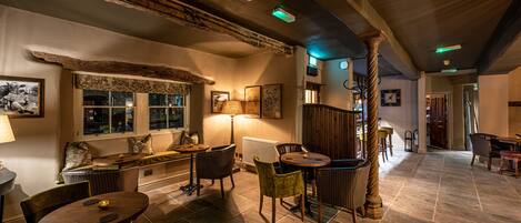 A cozy interior with a stone floor, wooden beams, a fireplace, and a dining area with chairs and a table.