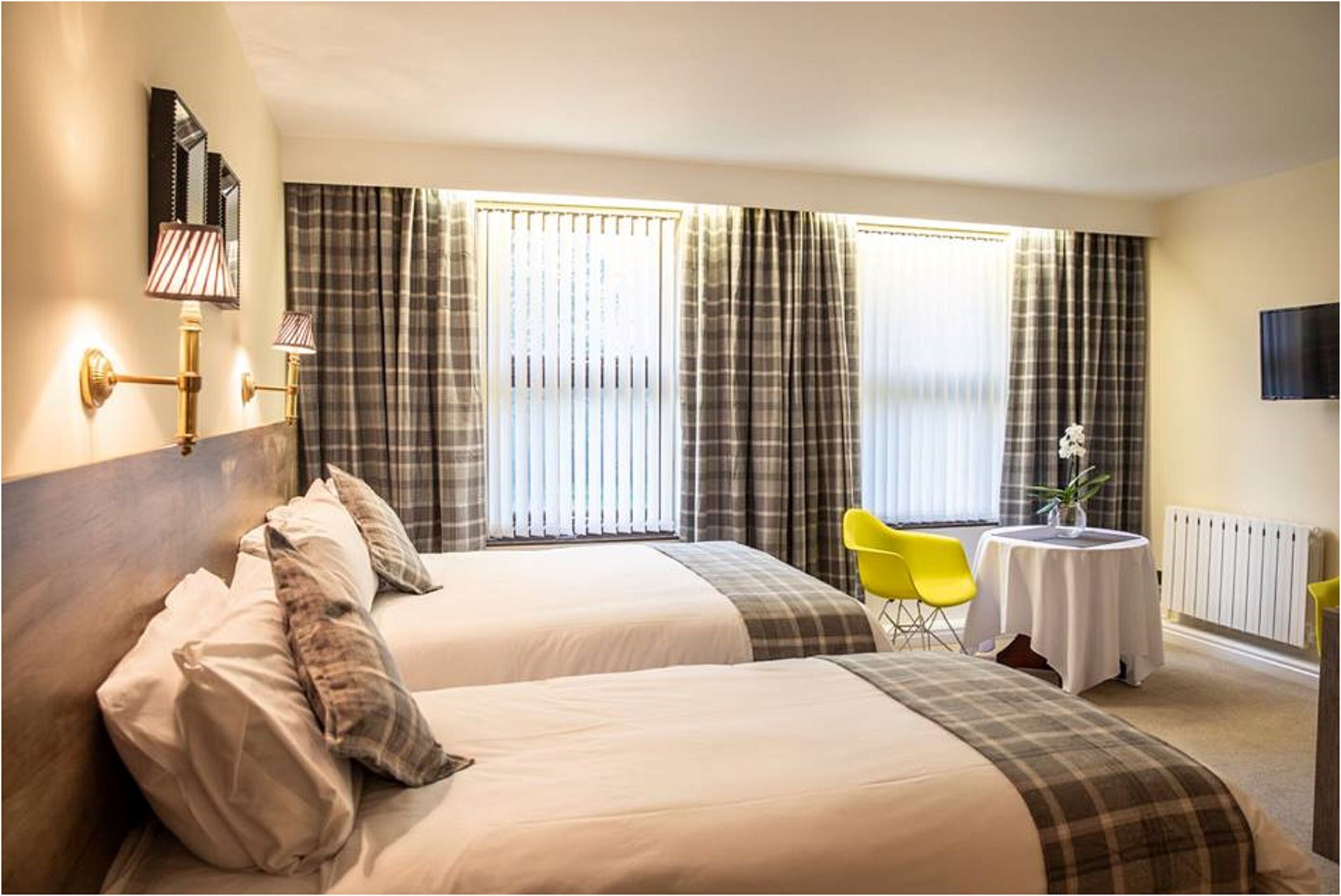 A hotel room with two beds, a small table with a vase, a yellow chair, and a TV on the wall.