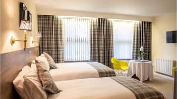 A hotel room with two beds, a small table with a vase, a yellow chair, and a TV on the wall.