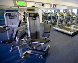 Uma academia com diversos equipamentos de ginástica, esteiras e uma televisão presa à parede.
