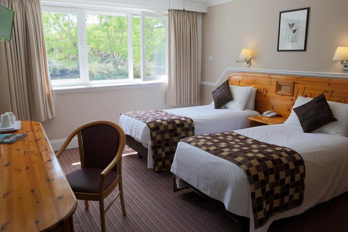 A hotel room with two beds, a wooden table, a chair, and a window with curtains.