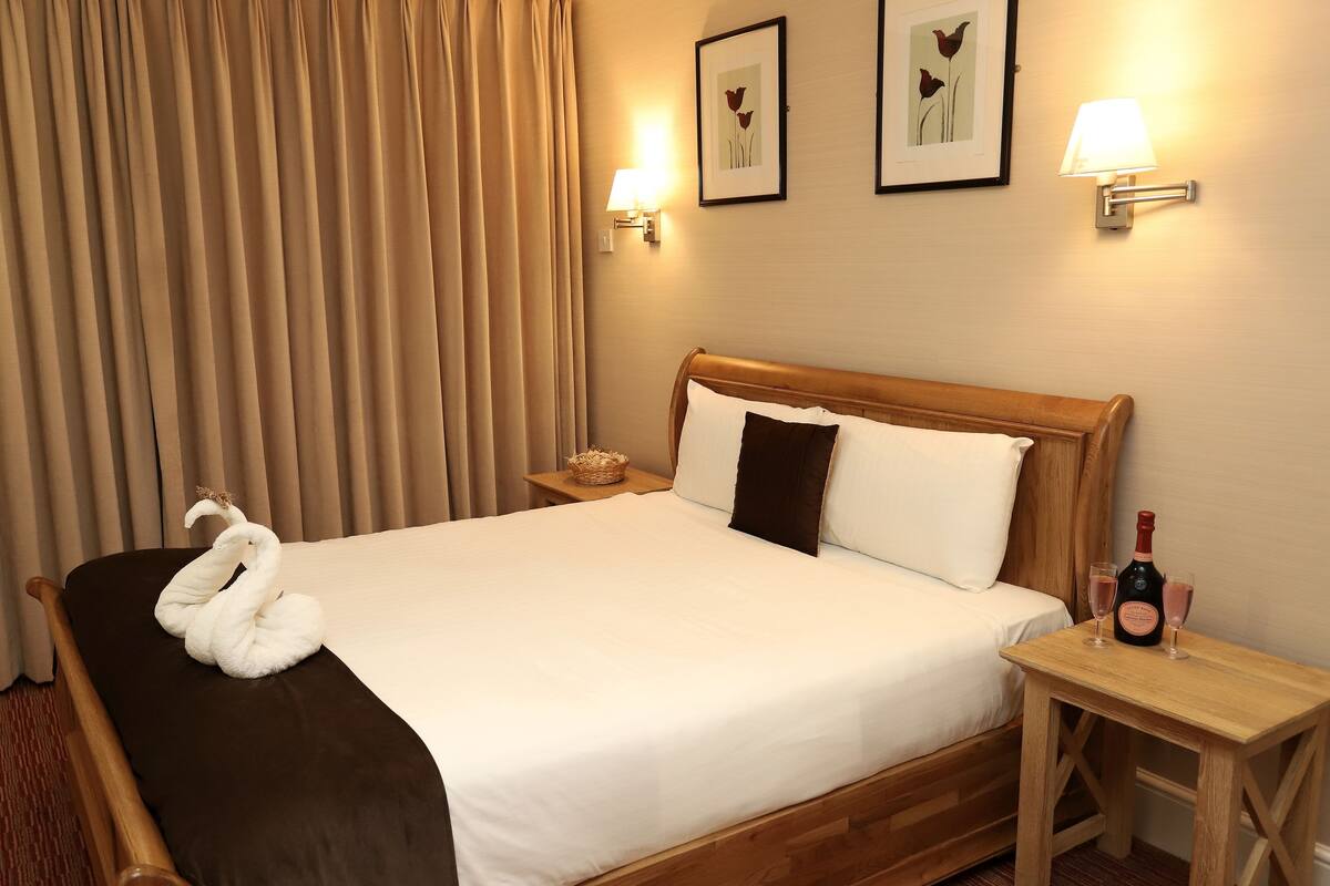 A hotel room with a large bed, a wooden headboard, a nightstand with a bottle and glasses, and a wall-mounted lamp.