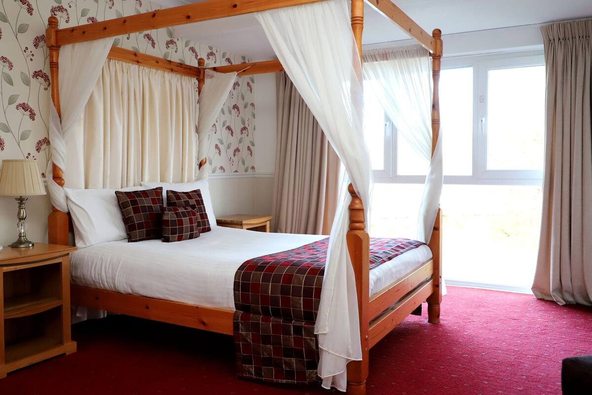 A four-poster bed with white bedding and a red and brown checkered throw. There is a nightstand with a lamp and a small table beside the bed. The room has floral wallpaper and a window with curtains.