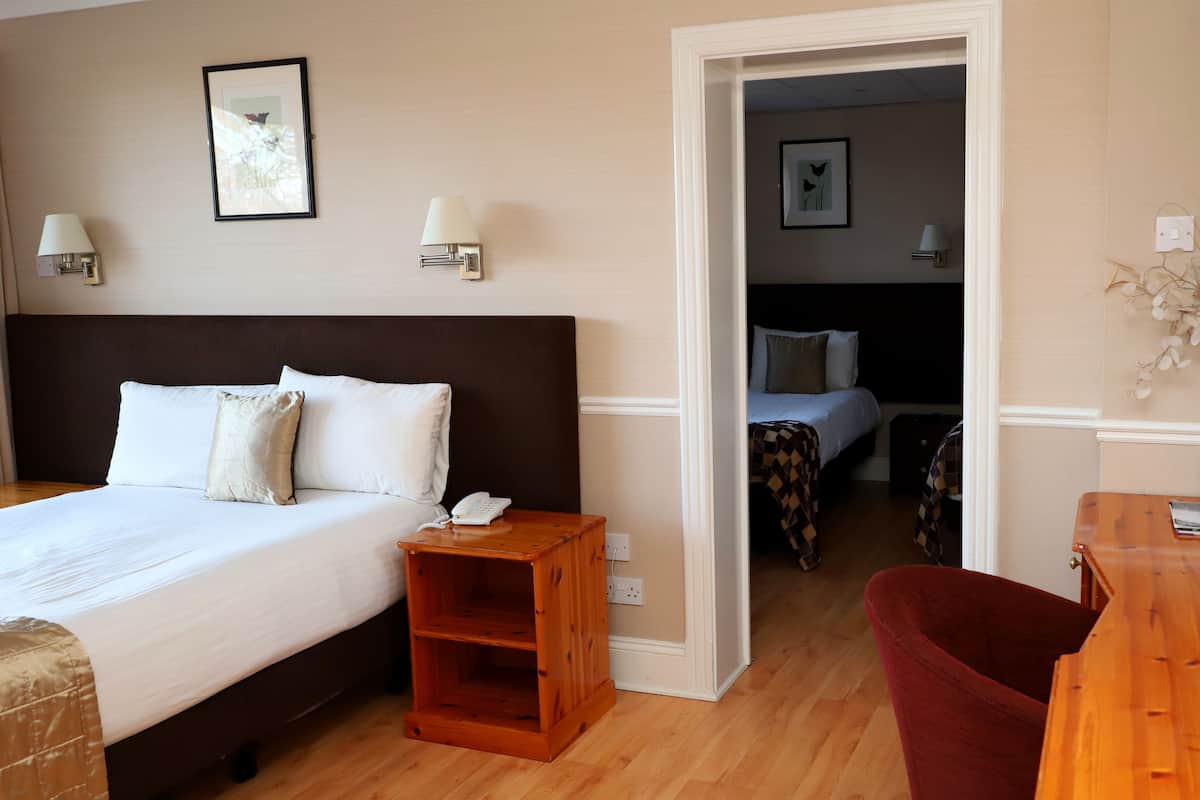 A hotel room with a large bed, bedside table, and a wooden nightstand. There is a chair and a small table with a vase of dried flowers.