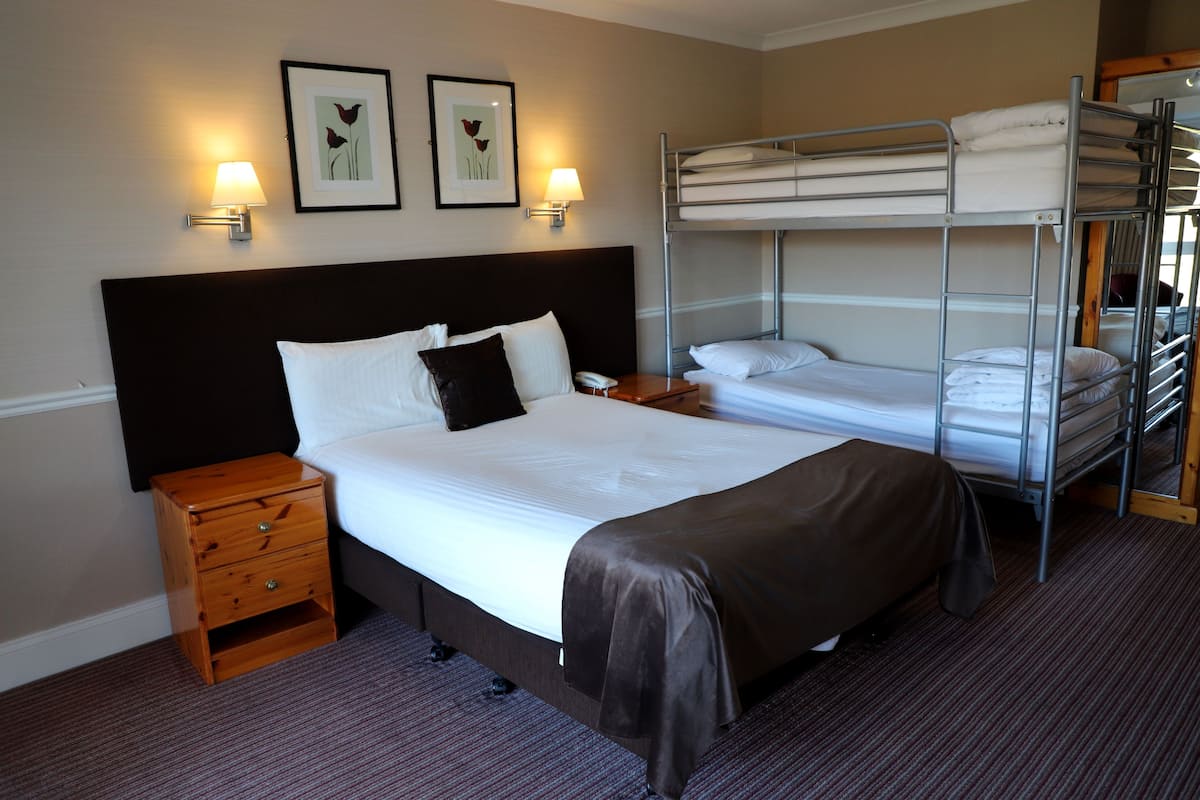 A hotel room with a single bed, a wooden nightstand, a bedside table, a lamp, and a bunk bed.