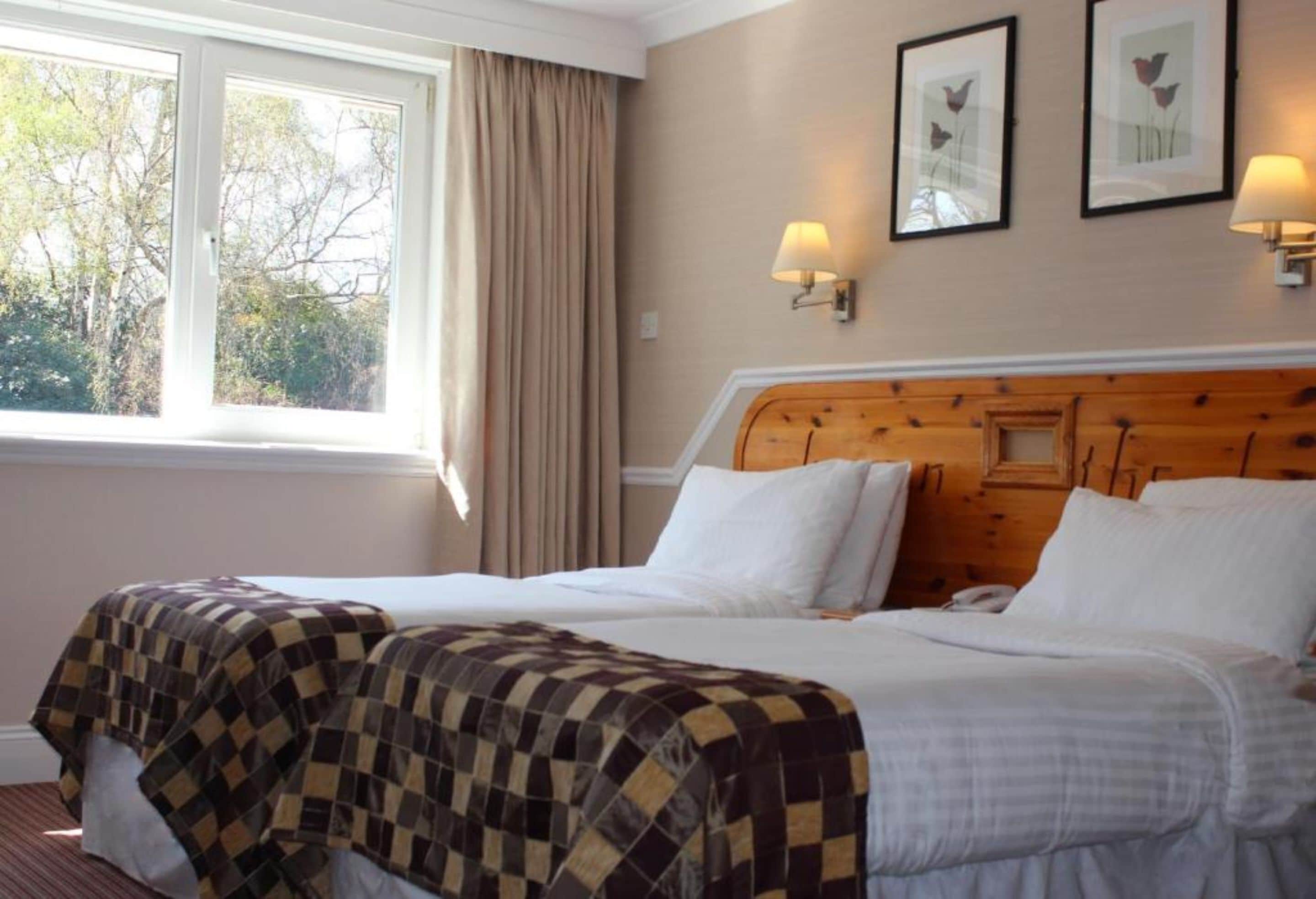 A hotel room with two beds, a large window with curtains, wall-mounted lamps, and framed floral artwork.