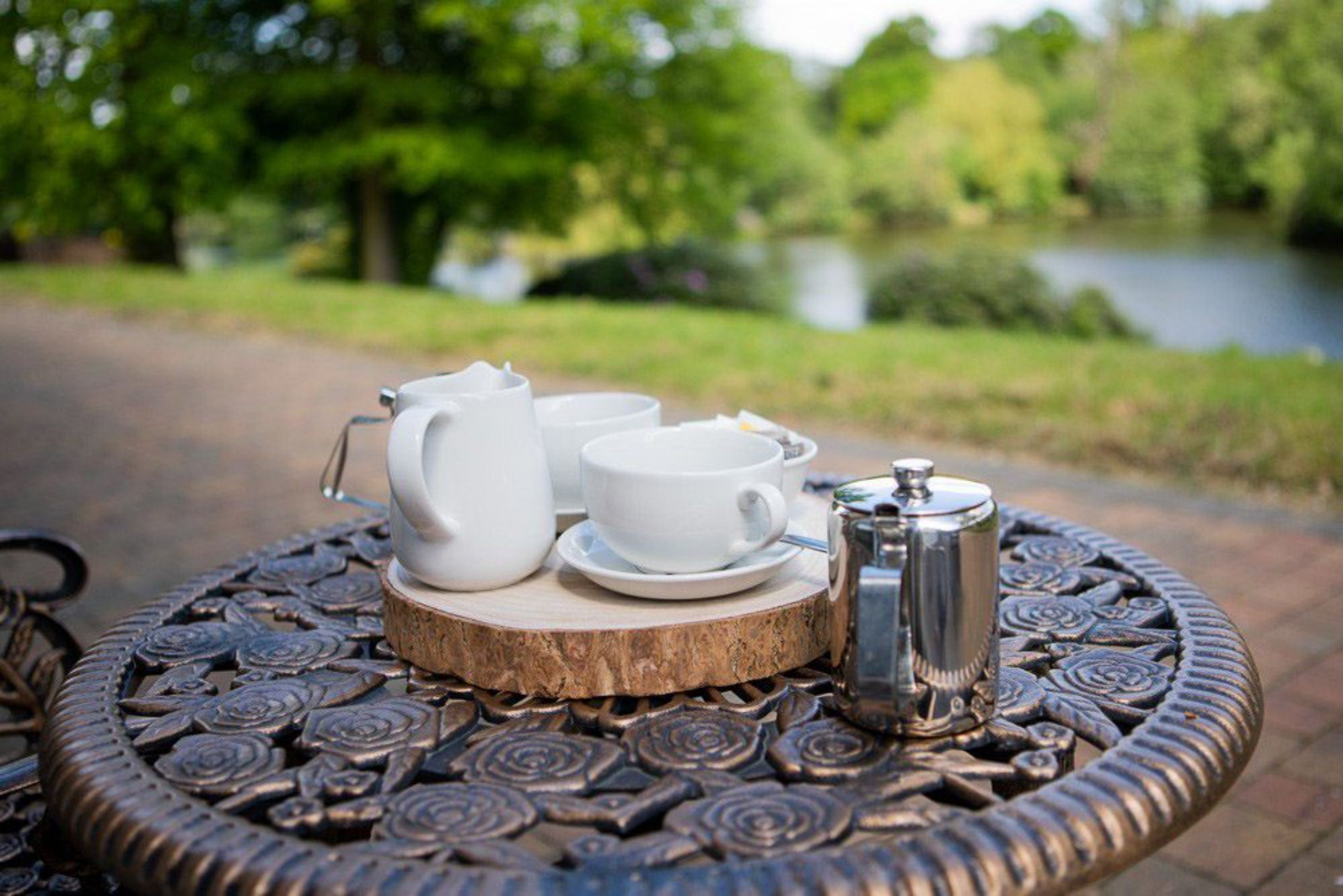 A metal table with a rustic wooden coaster, a white teapot, two white cups, and a silver coffee pot.