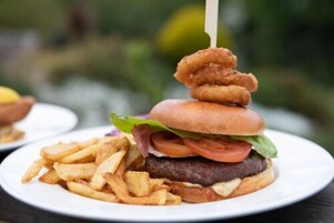 A burger with a stack of fried items on top, a side of fries, and a toothpick.