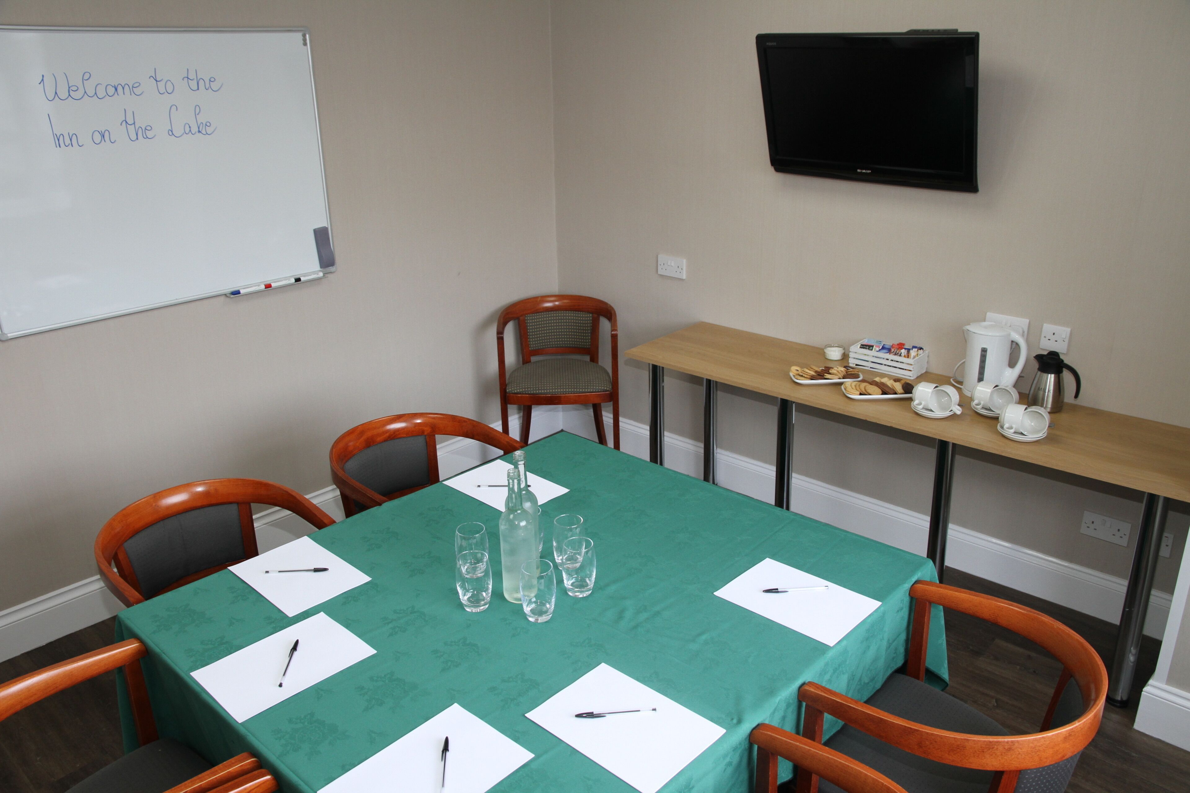 Un salón de conferencias con una pizarra, una mesa con mantel verde, sillas, una estación de café y un televisor en la pared.