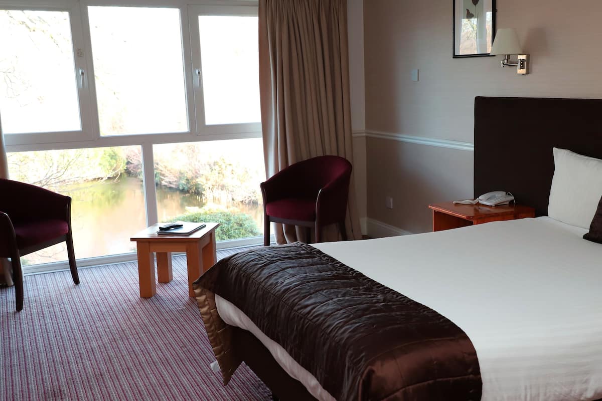 A hotel room with a large bed, a desk with a phone, a chair, and a view of a body of water through the window.