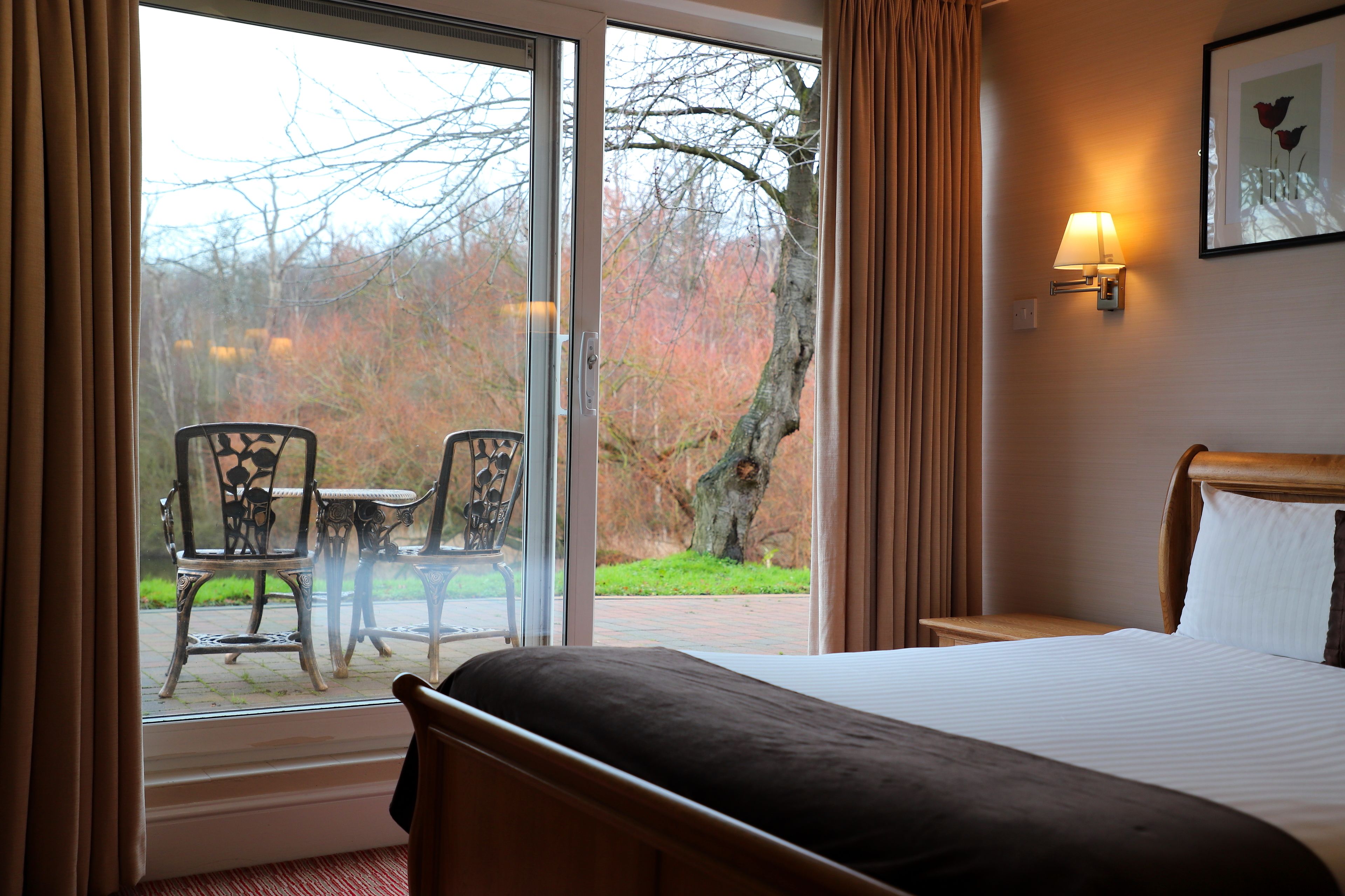 Une chambre d’hôtel avec un lit, une fenêtre avec des rideaux, une table avec des chaises et une lampe fixée au mur.