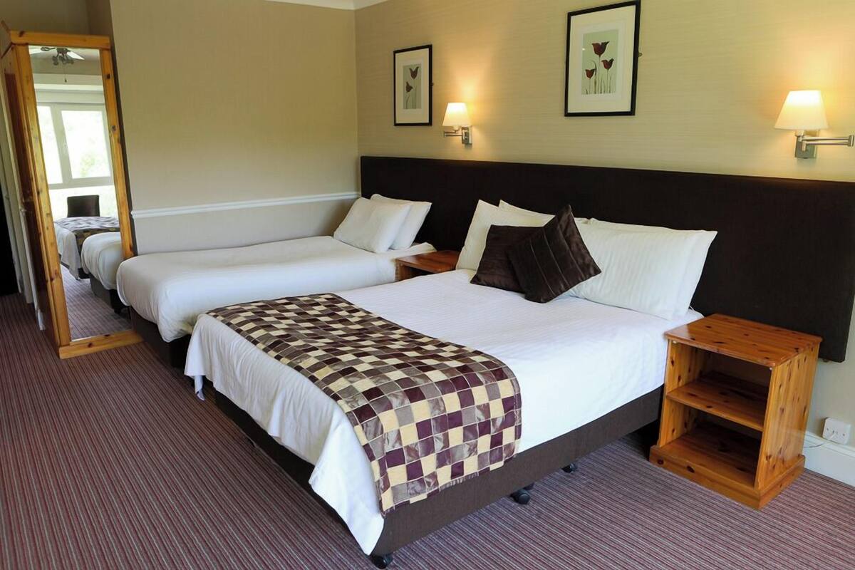A hotel room with two single beds, a large bed with a checkered blanket, a wooden nightstand, and a mirror.