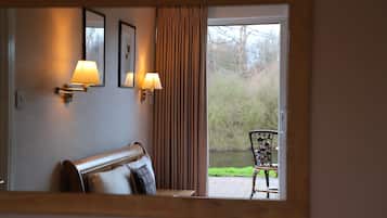 Ett hotellrum med en säng, två väggmonterade lampor, ett fönster med gardiner och utsikt över en trädgård samt en stol utomhus.