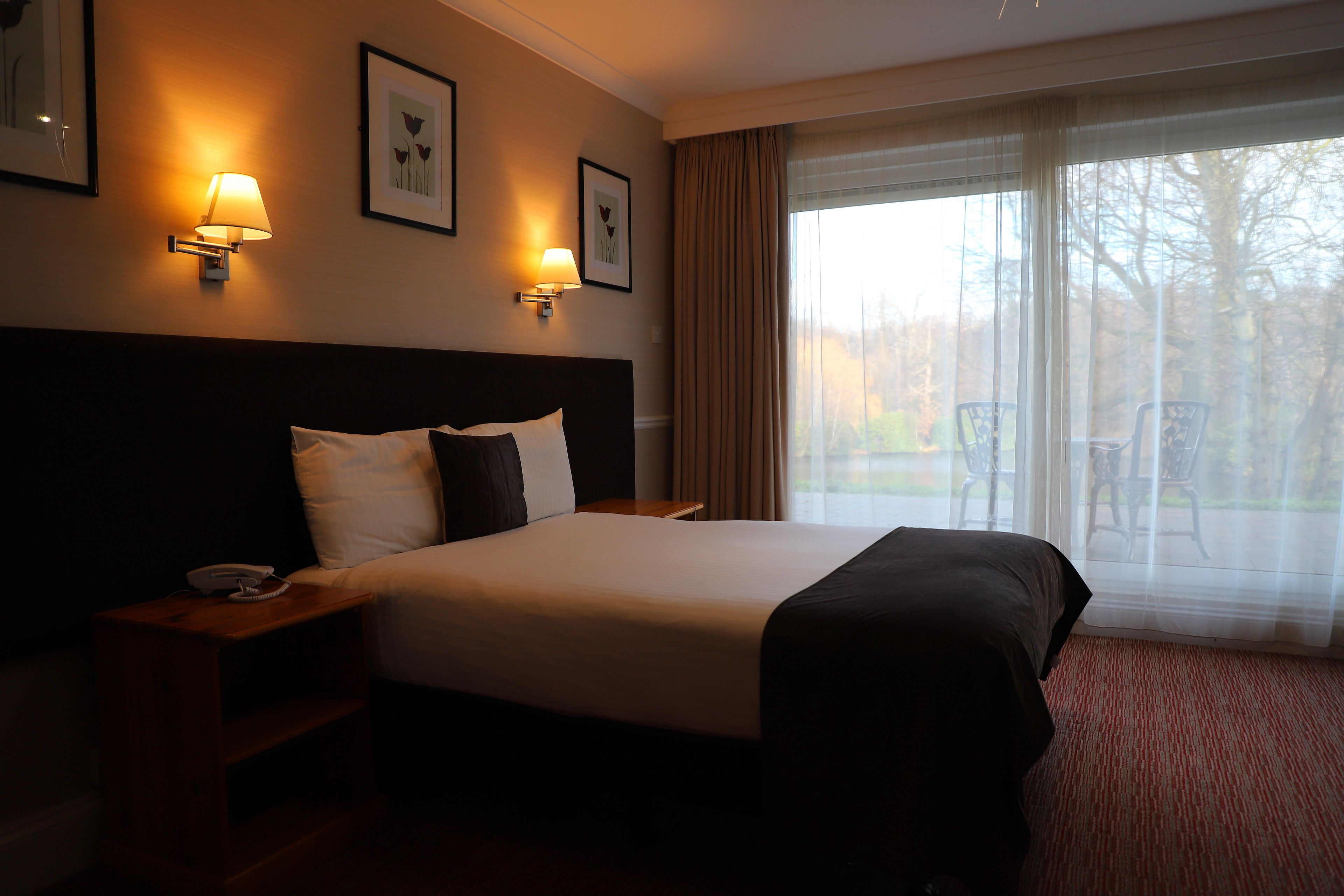 Habitación de hotel con una cama grande, dos lámparas de noche, una mesita de noche de madera y vistas a sillas exteriores a través de cortinas translúcidas.