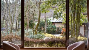 Quarto com uma grande janela que oferece vista para um jardim e um edifício tradicional. Há uma pequena mesa e duas cadeiras no quarto.