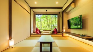 Um quarto japonês tradicional com tatames, uma mesa baixa, almofadas, uma TV e uma janela com vista para árvores.