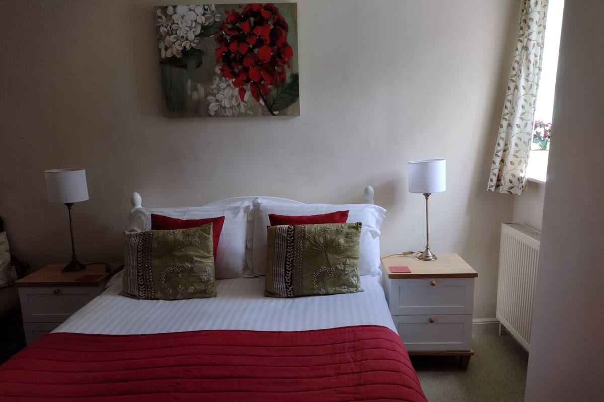 Una cama bien hecha con una manta roja, almohadas blancas y un cuadro floral encima. Hay dos mesitas de noche con lámparas y un radiador en la esquina.