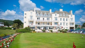 A large, multi-story hotel with a well-maintained lawn in front, multiple balconies, and a parking area. - The Belmont (Sidmouth)