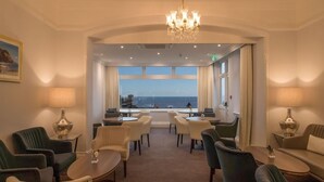 A hotel lobby with a view of the sea, furnished with chairs and tables, and a chandelier. - The Belmont (Sidmouth)