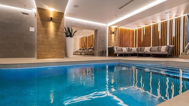 A modern indoor swimming pool with a clear blue water surface, surrounded by a lounge area with seating and a wooden wall design. - The Belmont (Sidmouth)