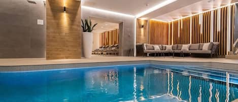 Piscina interior moderna con agua azul cristalina, zona de estar con sofá y pared de madera con listones verticales.