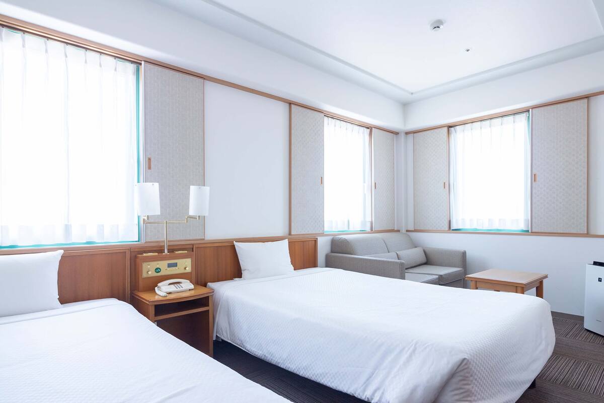 Habitación de hotel con dos camas, una mesita de noche con teléfono, una pequeña mesa y una zona de estar con un banco.