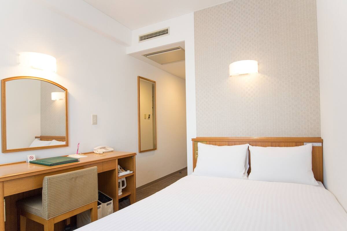Habitación de hotel con una cama, un escritorio con teléfono, una silla, un espejo y una lámpara en la pared.