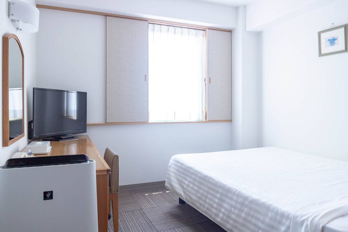 Habitación de hotel con cama, escritorio con televisor, silla, refrigerador pequeño y ventana con persianas.