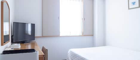 A hotel room with a bed, a desk with a television, a chair, a small refrigerator, and a window with blinds.