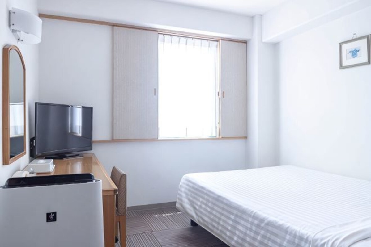 Una habitación de hotel con una cama, un escritorio con televisor, un espejo y una ventana con persianas.